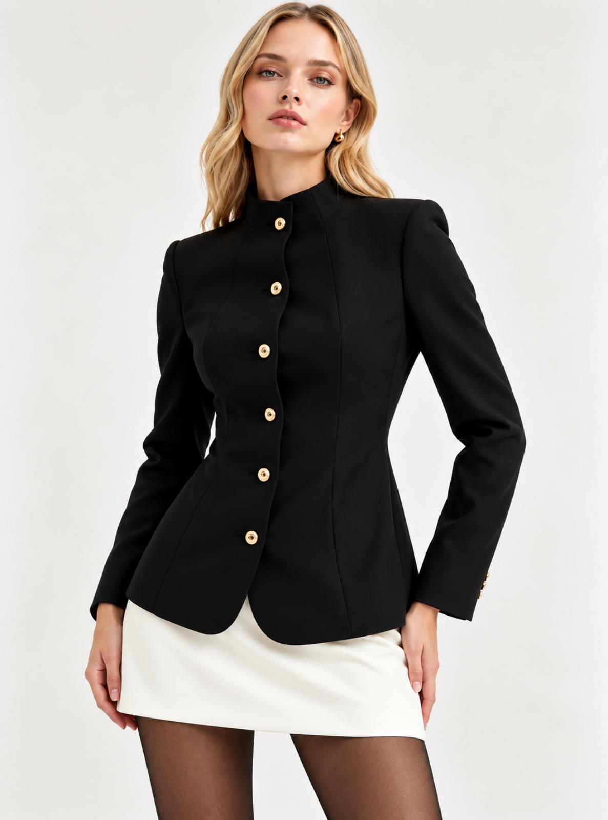 A woman modeling a structured black blazer with gold buttons and a high stand collar, paired with a white mini skirt and sheer black tights, against a light gray studio background.