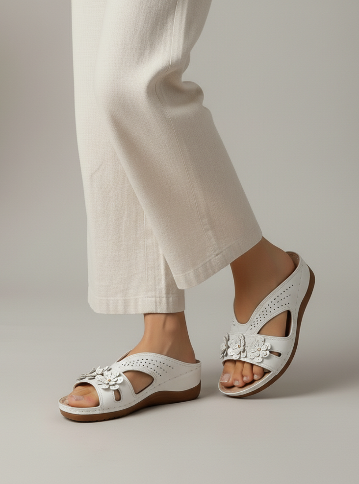 White sandals with floral details worn by a person in light-colored pants on a neutral background