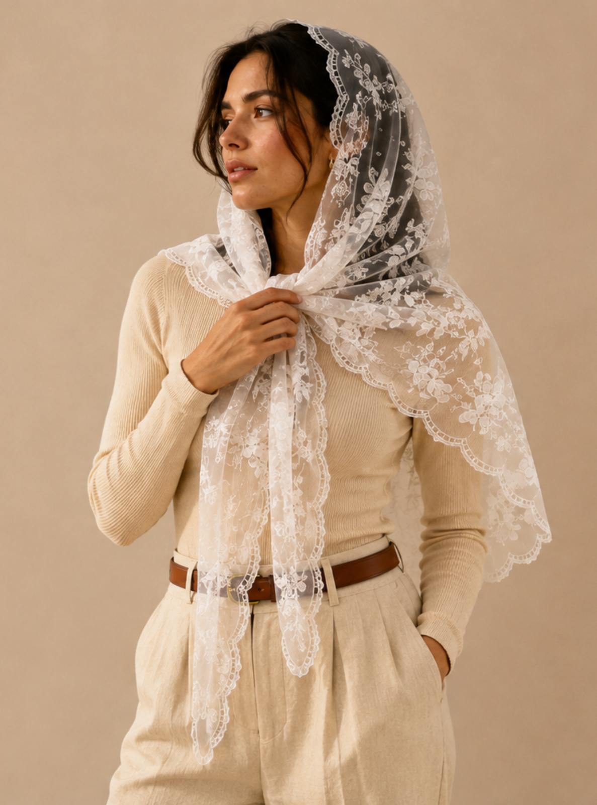 A woman wearing a cream lace headscarf and matching beige outfit, standing against a warm beige background with soft window light shadows.