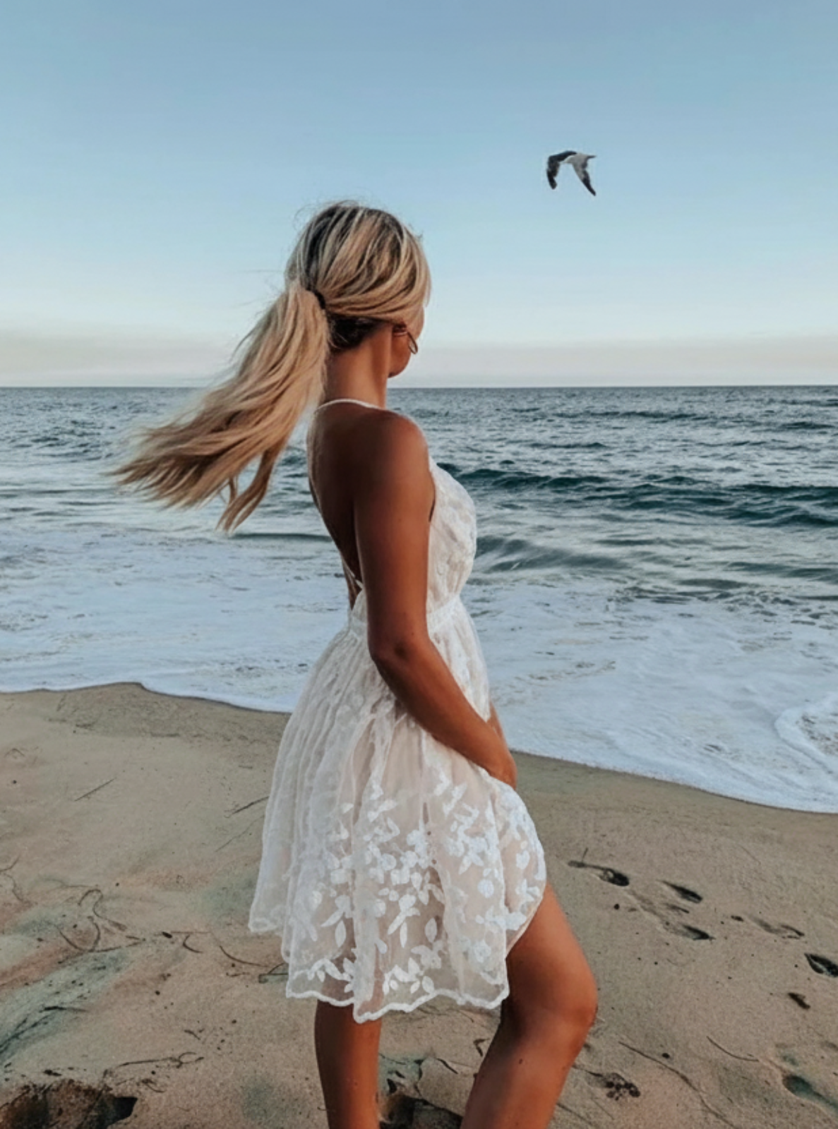 Woman in a white dress standing on a beach with ocean waves and a clear sky.