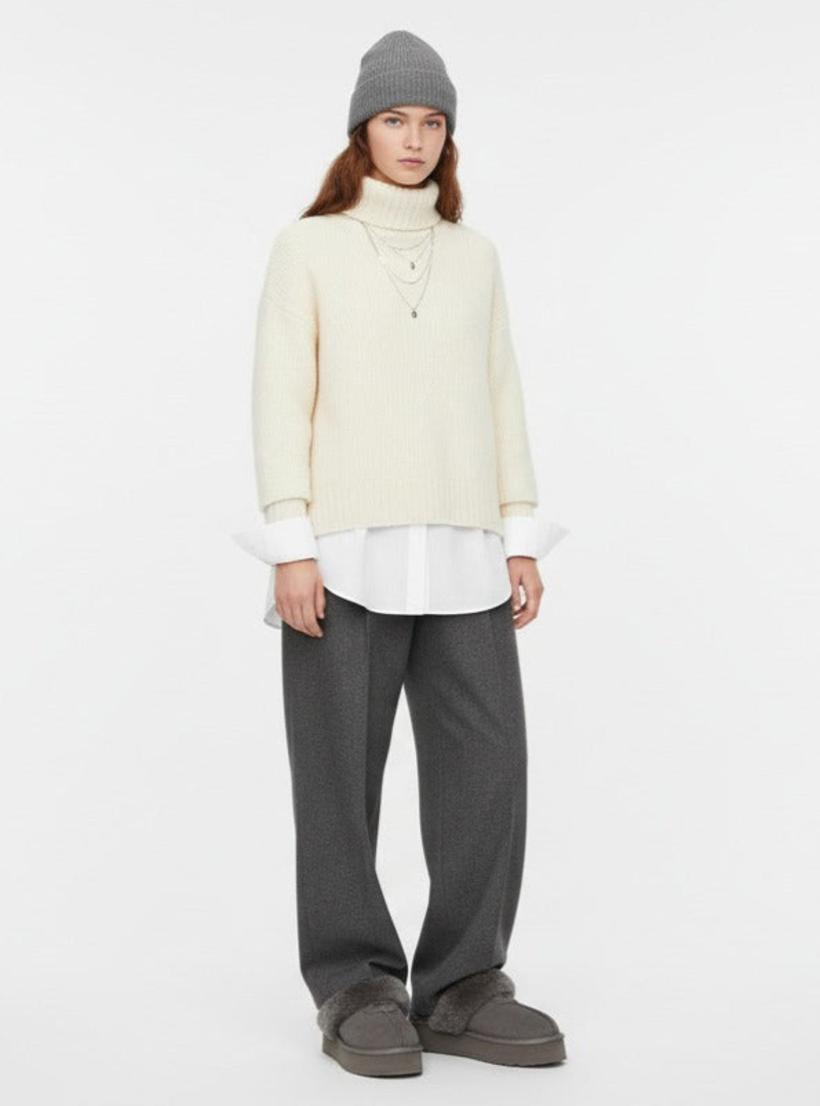 Person wearing a cream sweater, white shirt, gray pants, and gray beanie on a white background