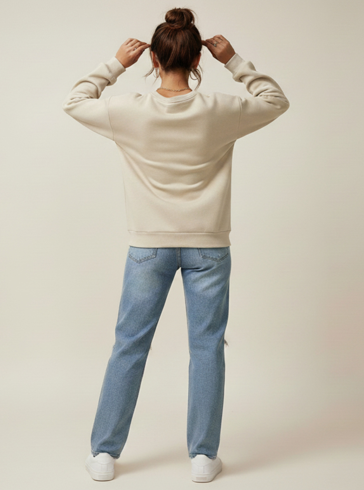 Person wearing a beige sweater and blue jeans standing against a plain background