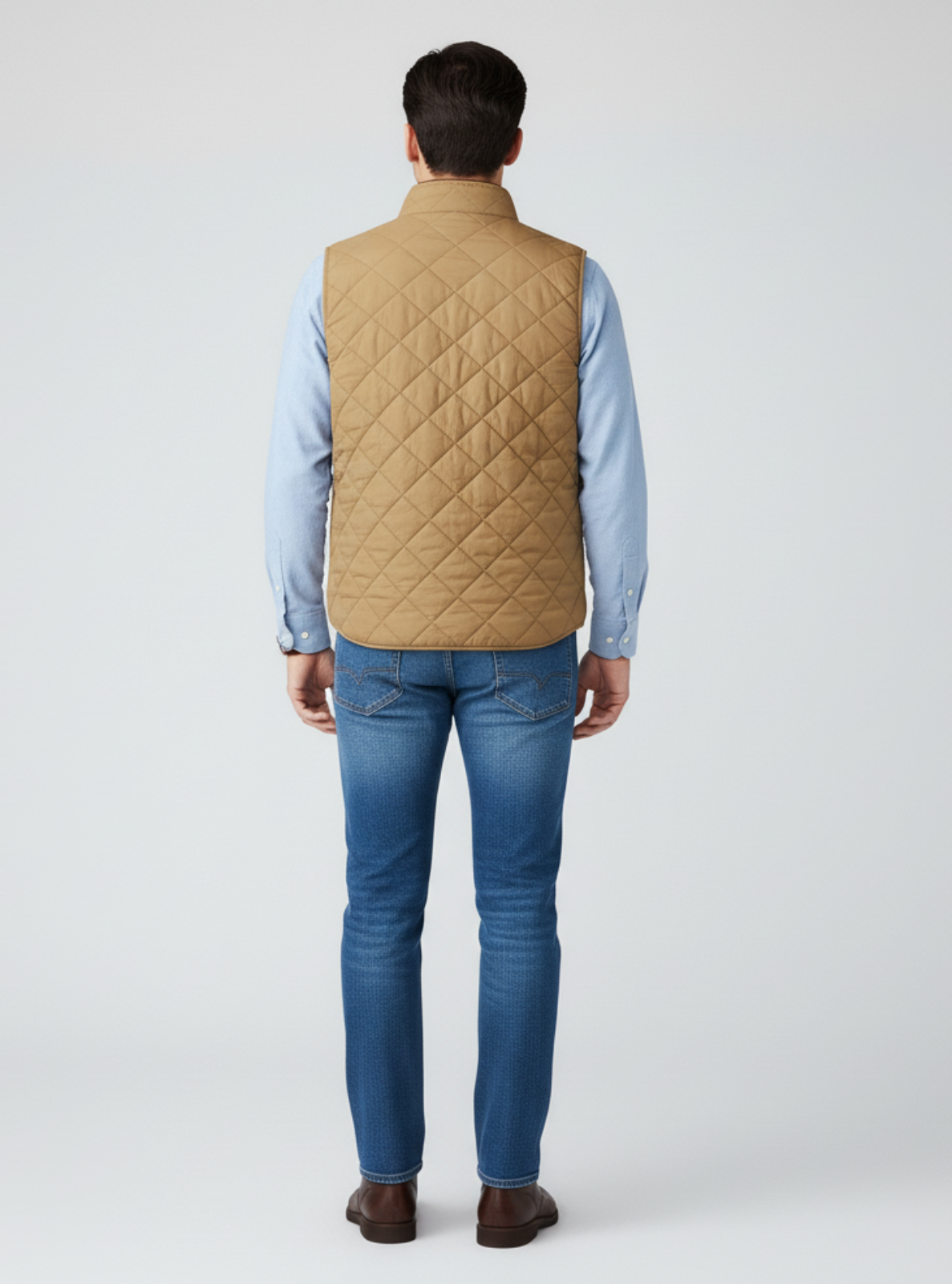 Man wearing a beige quilted vest over a light blue shirt and blue jeans on a white background