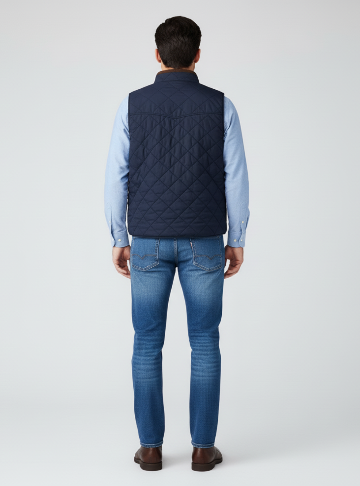 Man wearing a navy quilted vest over a light blue shirt and blue jeans on a white background