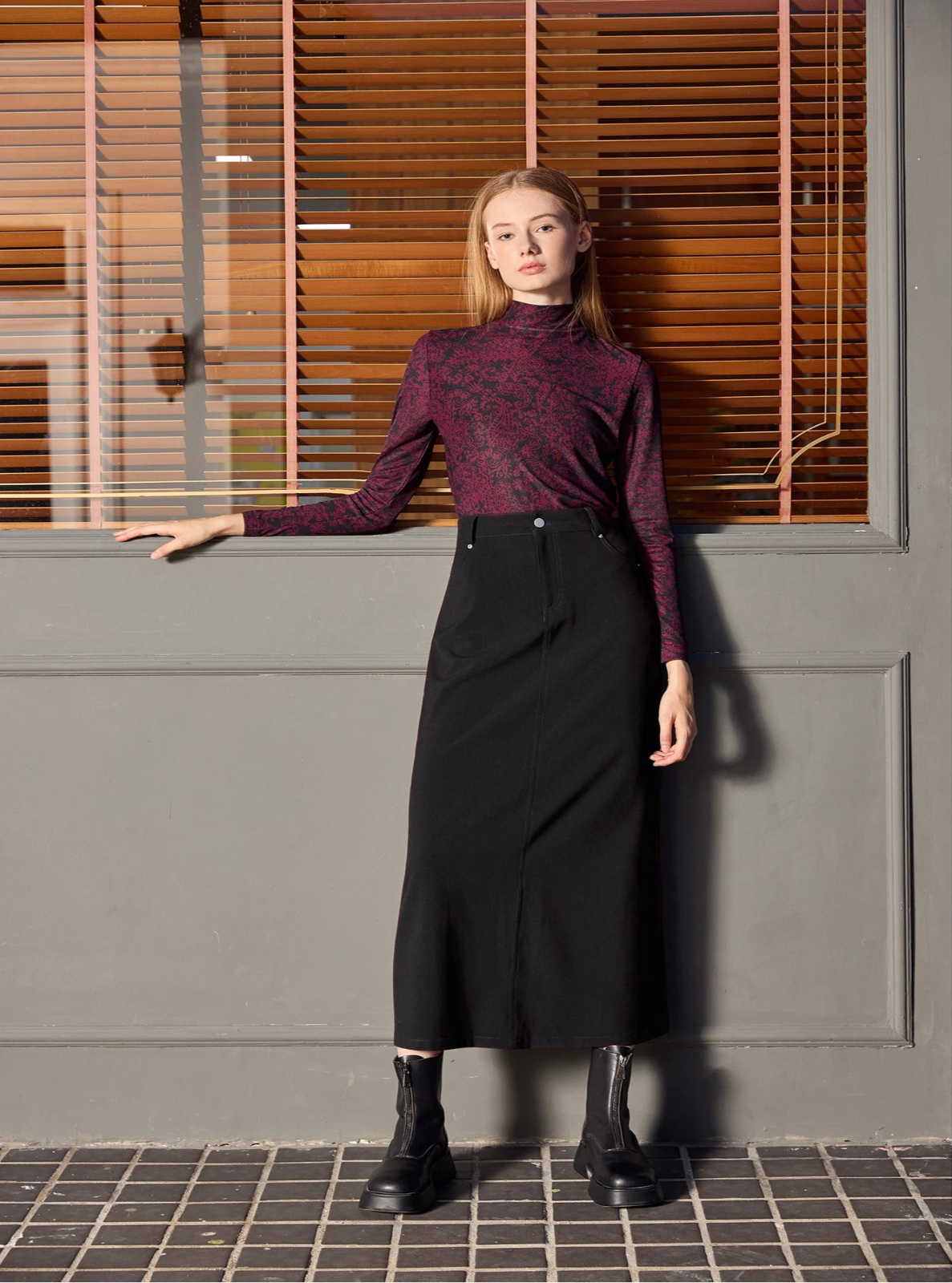 Woman wearing a maroon patterned top and black skirt standing against a gray wall with wooden blinds.