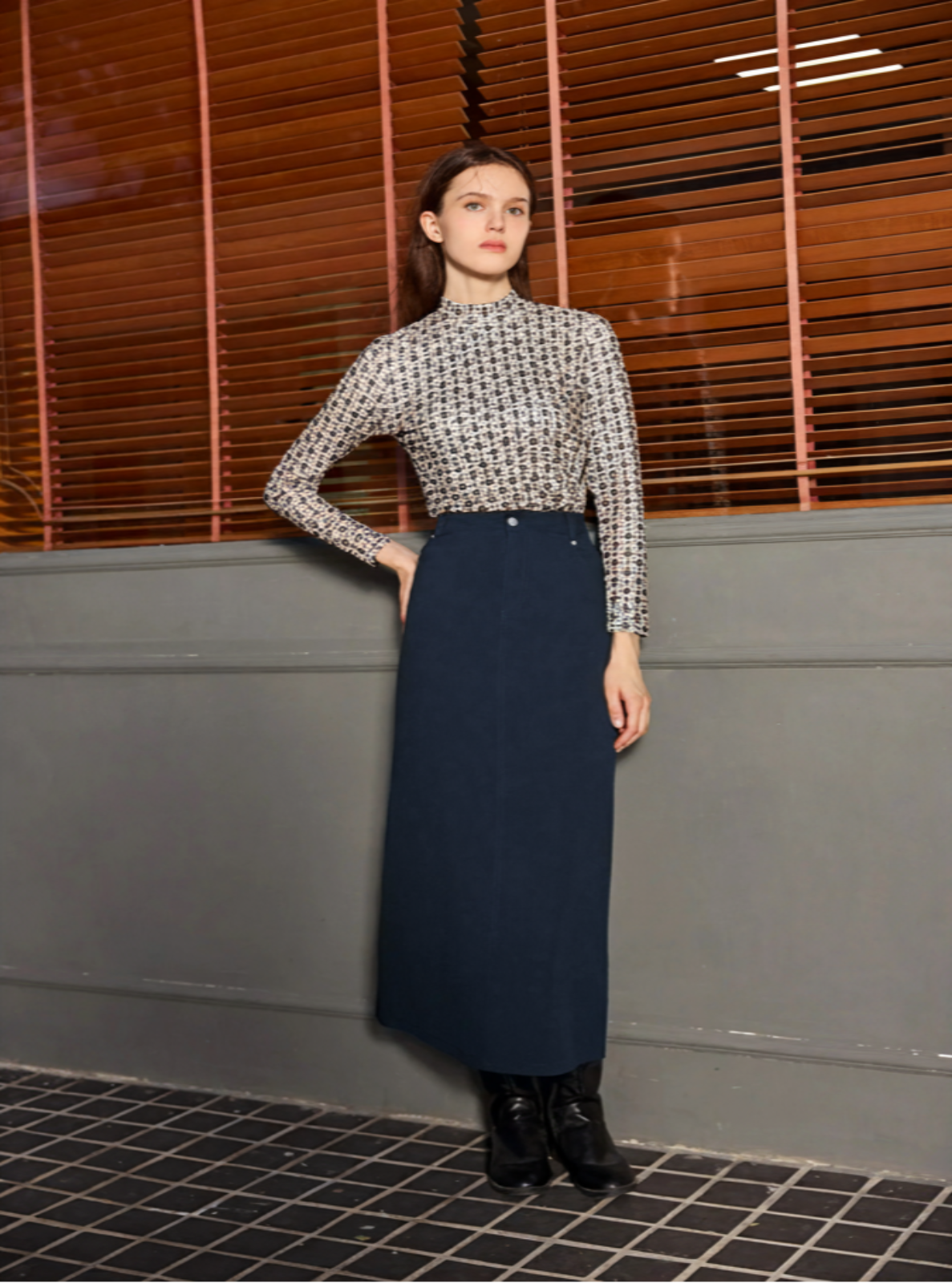 Woman wearing a patterned blouse and navy skirt standing against a neutral wall.