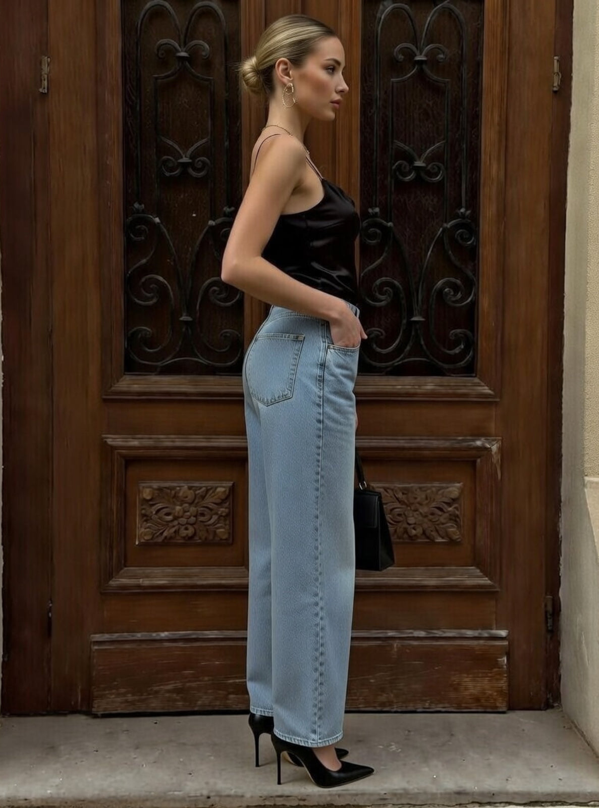 Woman in black top and light blue jeans standing in front of a wooden door.