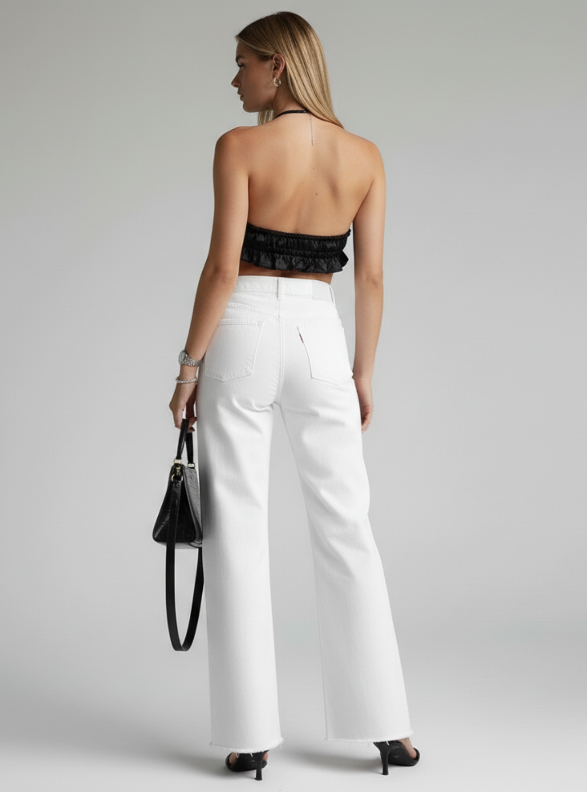 Woman wearing a black lace top and white pants, holding a black handbag on a gray background