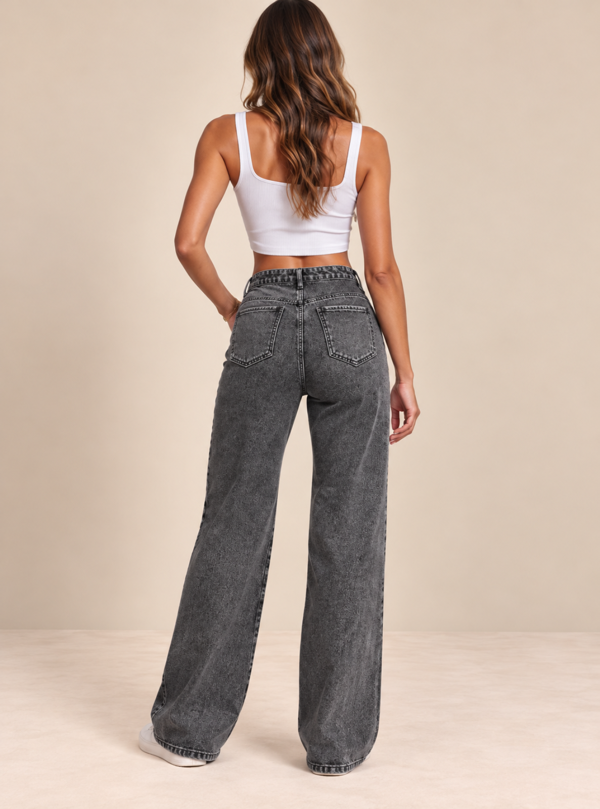 A woman viewed from behind wearing high-waisted, wide-leg gray denim jeans and a white crop top, standing against a solid beige studio background.