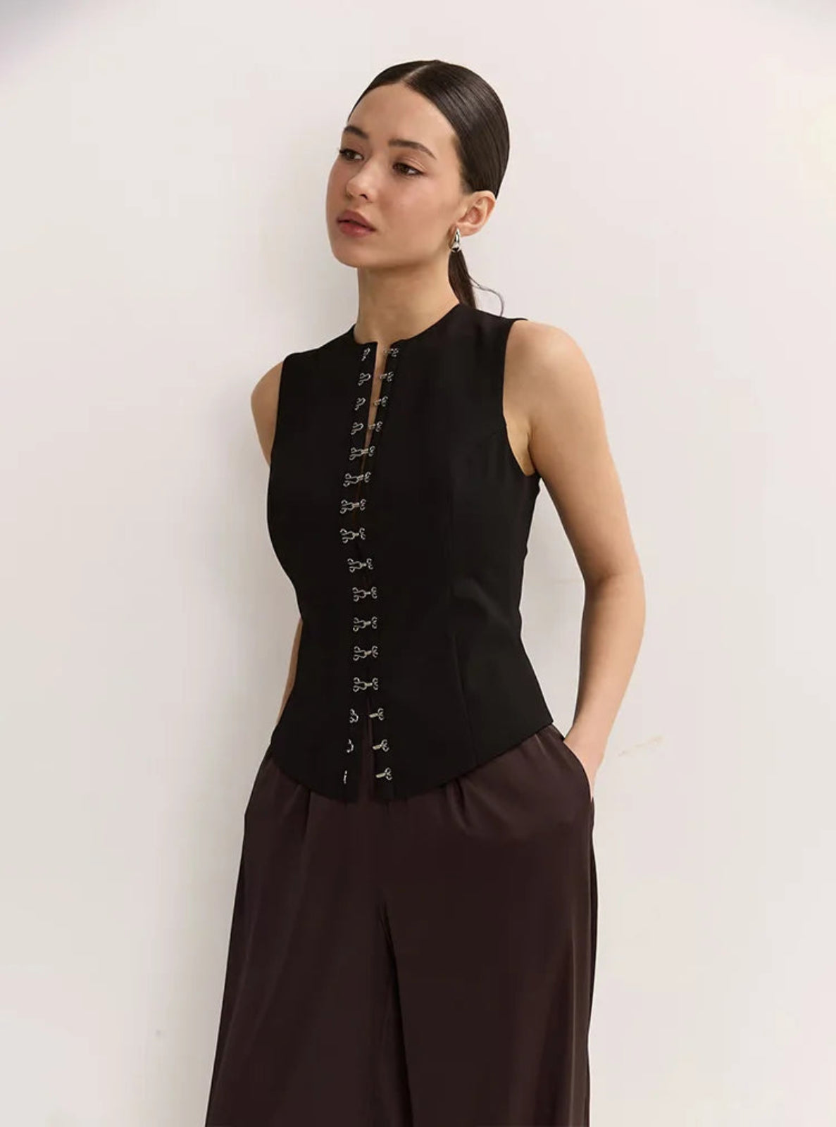 Woman wearing a black sleeveless top with lace-up front and brown pants against a white background
