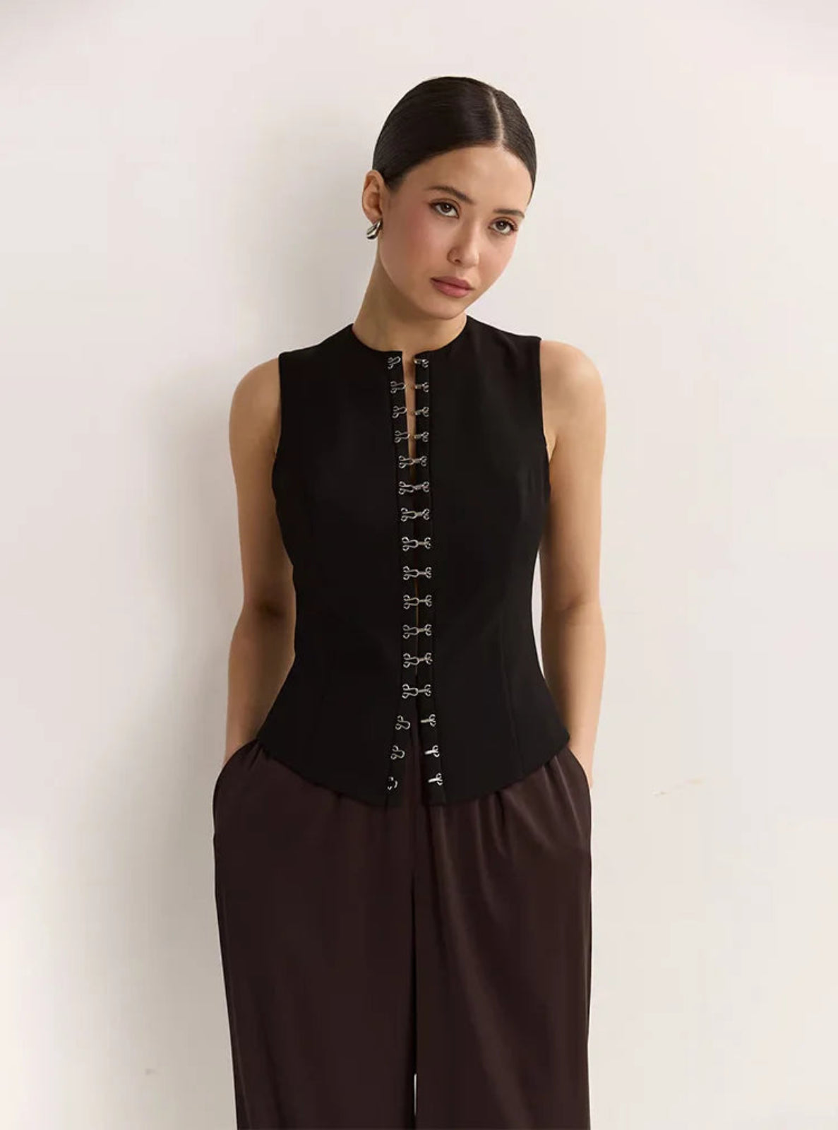 Woman wearing a black sleeveless top with decorative buttons and brown pants against a light background