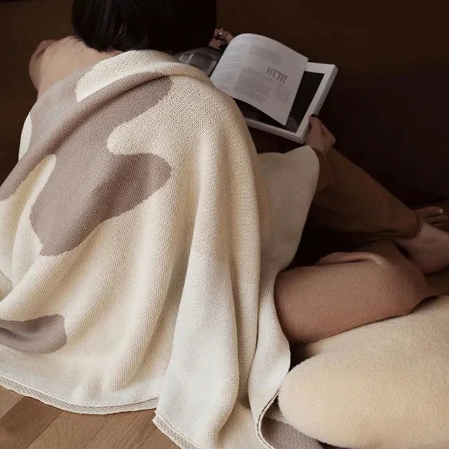 Person wrapped in a beige blanket reading a book on a wooden floor.