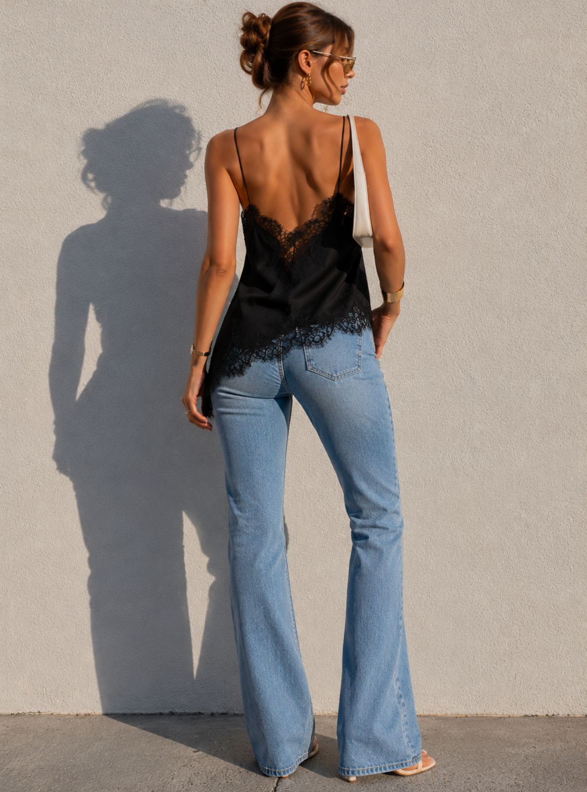 Woman wearing a black lace top and blue flared jeans against a plain wall.