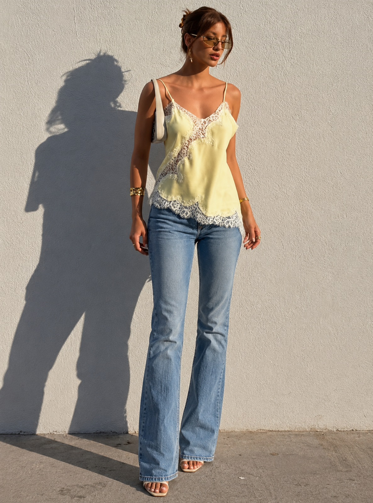 Woman wearing a light yellow lace top and blue jeans standing against a plain wall.