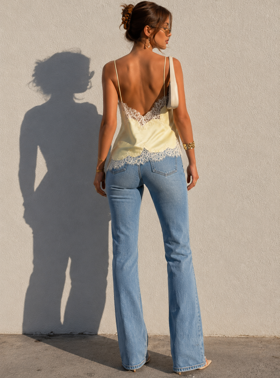 Woman wearing a yellow lace top and blue jeans standing against a plain wall with her shadow cast on the ground.