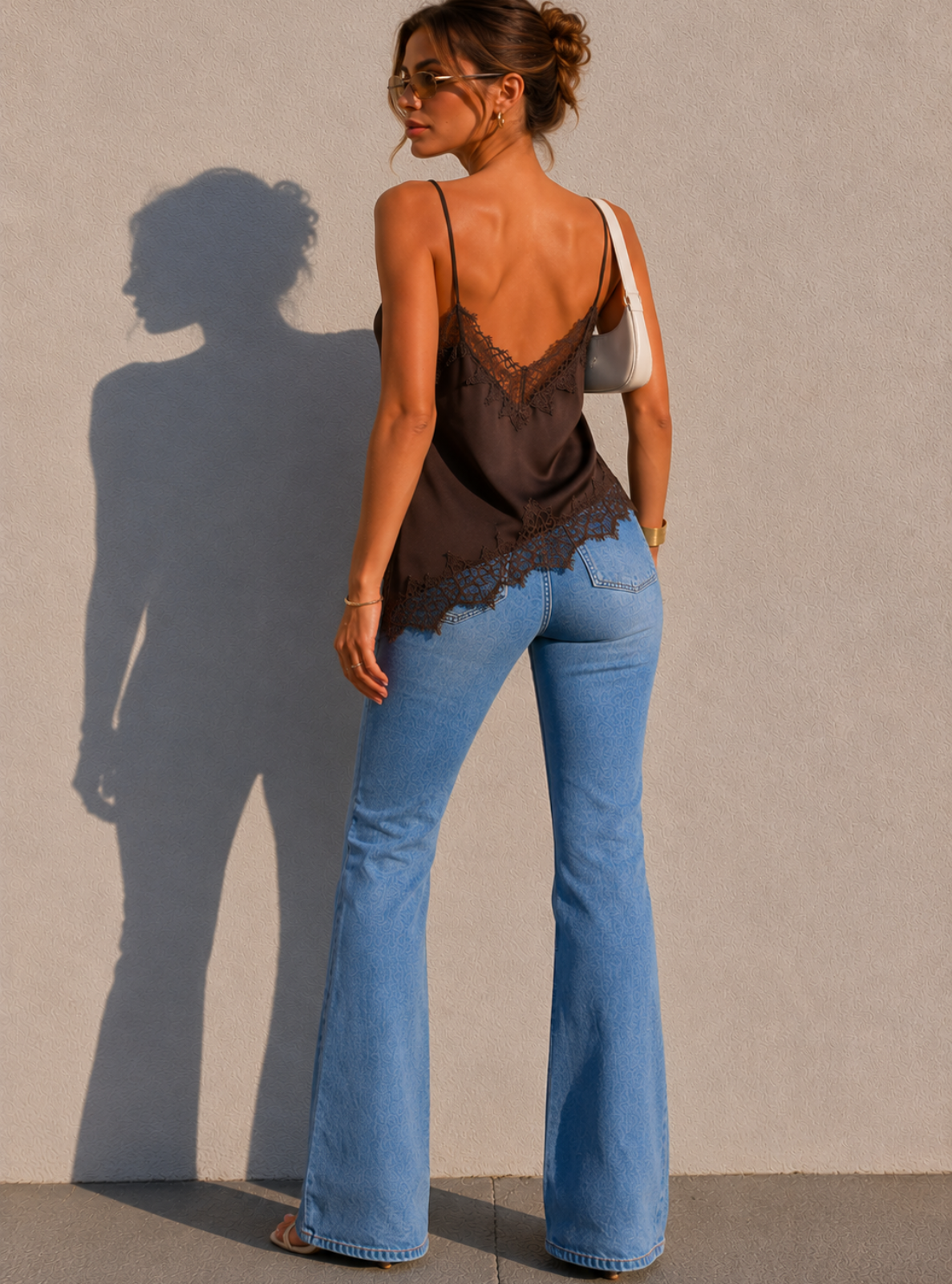 Woman wearing a black lace top and blue jeans standing against a plain wall with a shadow cast on the ground.