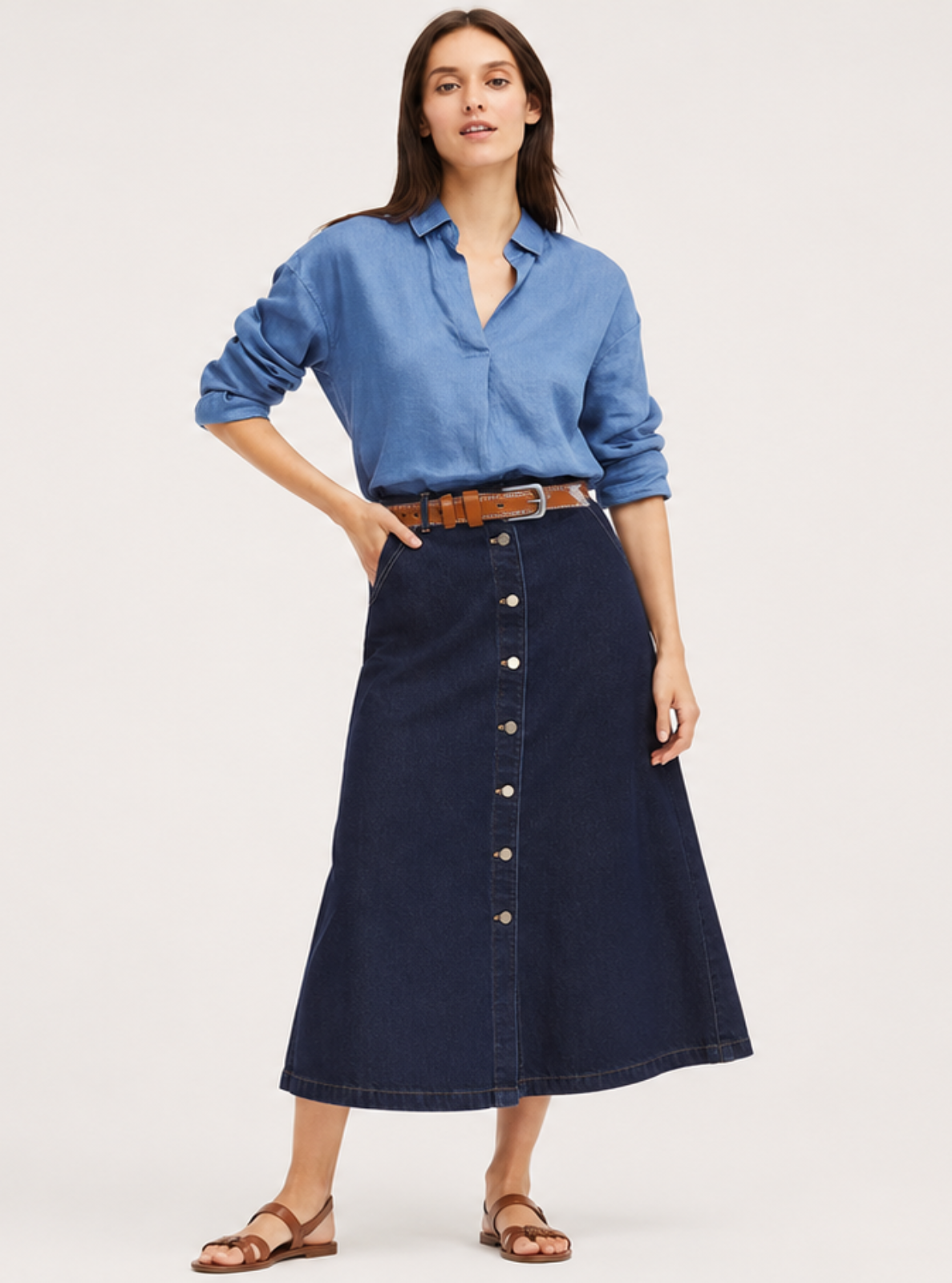 A woman modeling a dark navy midi button-front skirt with a brown belt, paired with a light blue button-up shirt and brown sandals, against a plain light gray studio background.