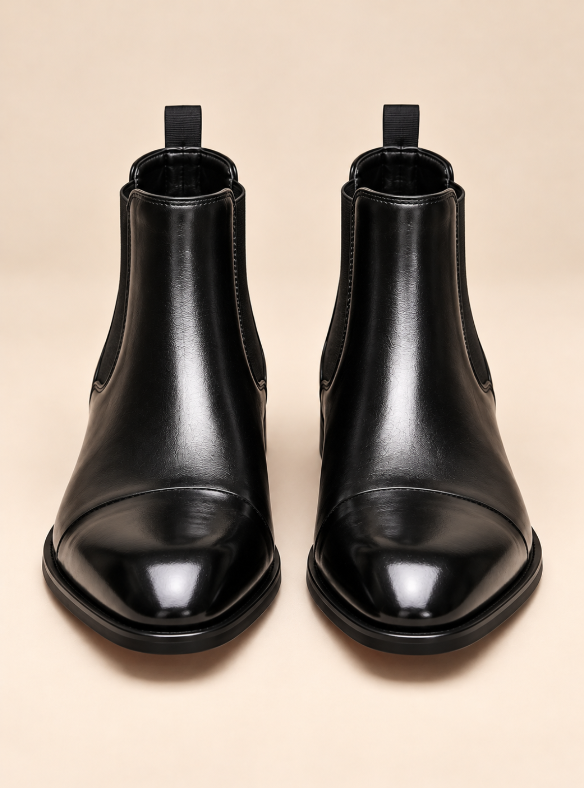 A pair of black leather Chelsea boots with a cap toe, shown from a front-facing overhead perspective on a solid beige background.