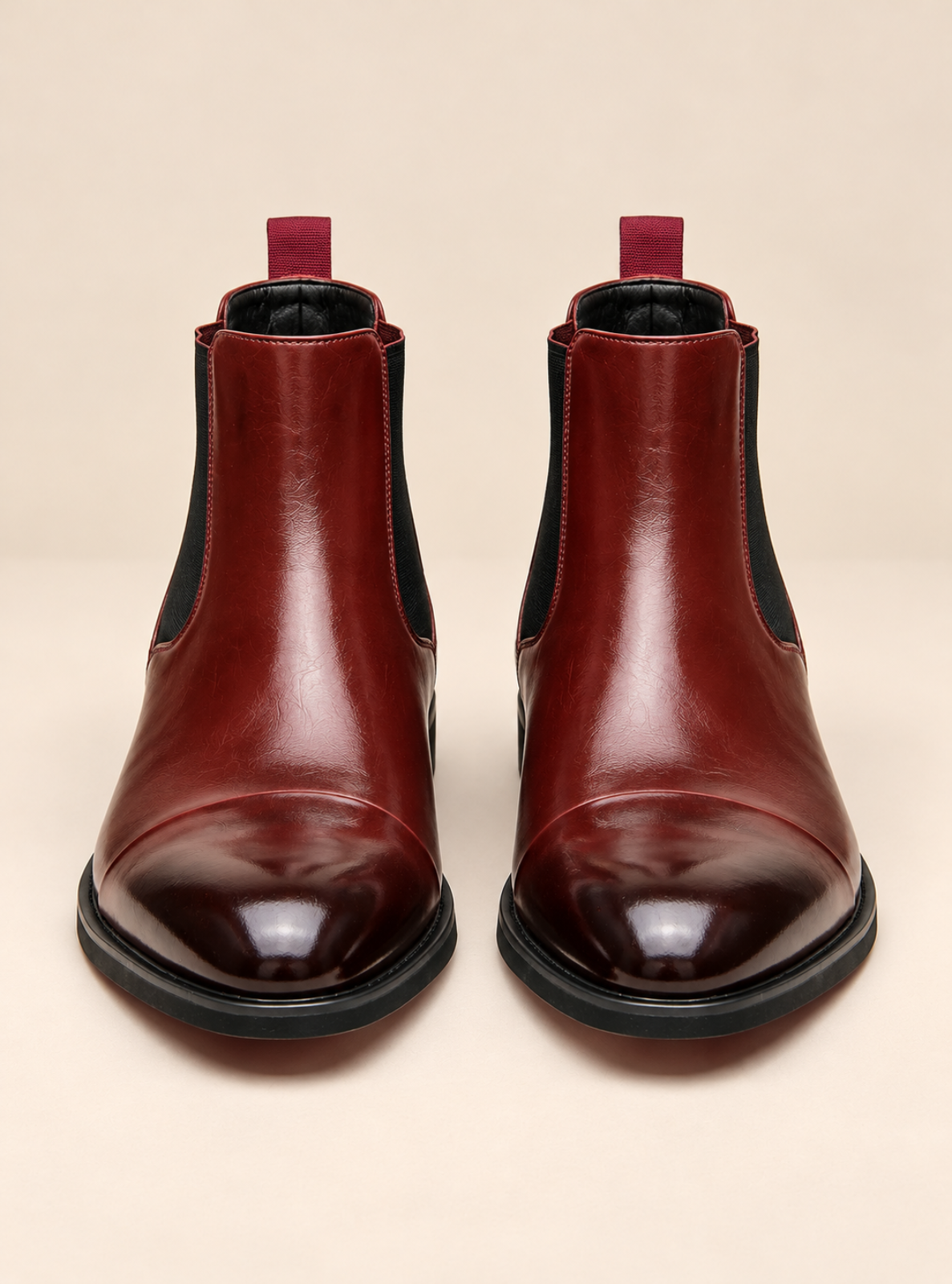 A pair of glossy burgundy leather Chelsea boots with black elastic side panels and black soles, shown from the front on a light beige background.