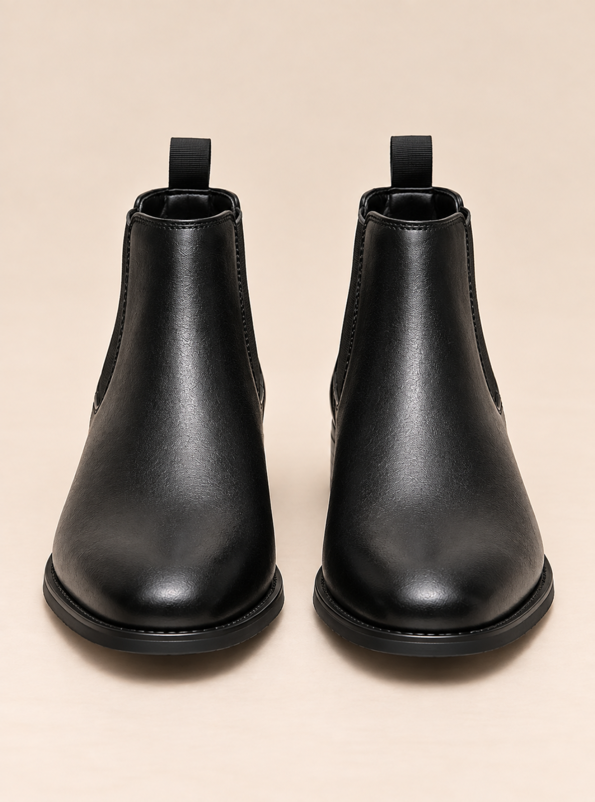 A pair of black leather Chelsea boots with elastic side panels and pull tabs, shown from a front-facing perspective on a light beige background.