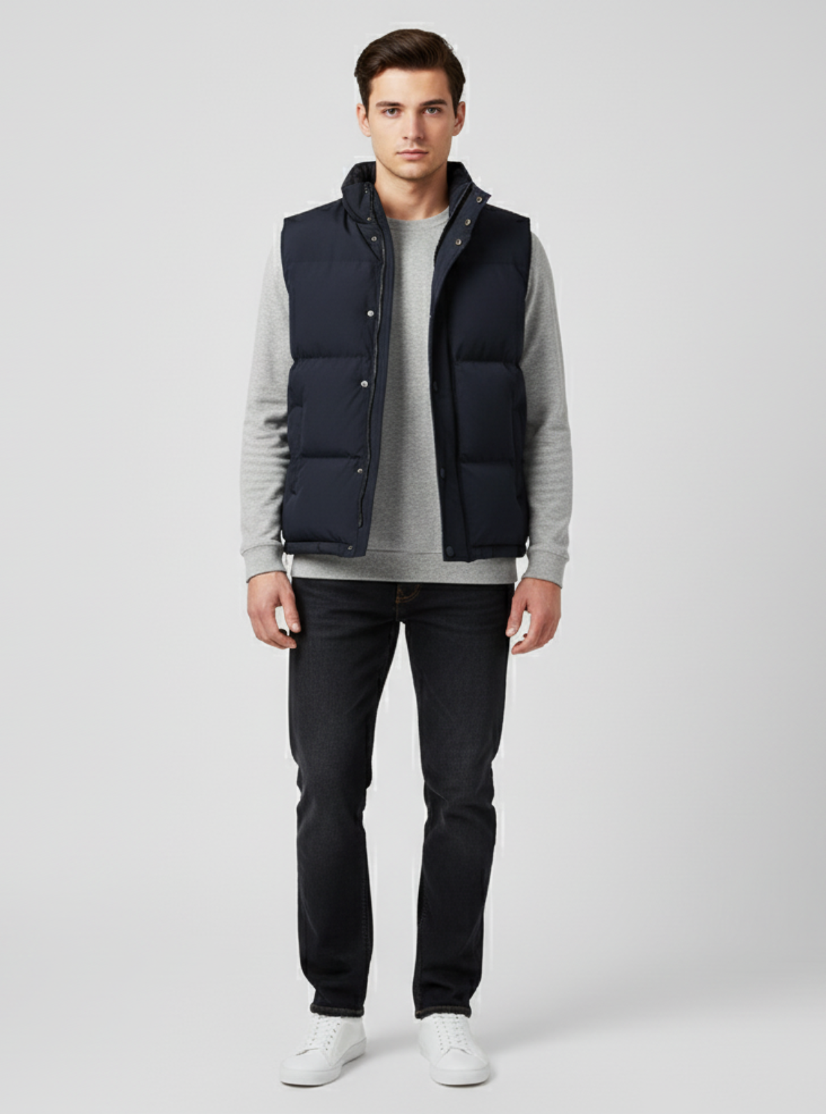 Man wearing a navy puffer vest over a gray sweater with black pants on a white background