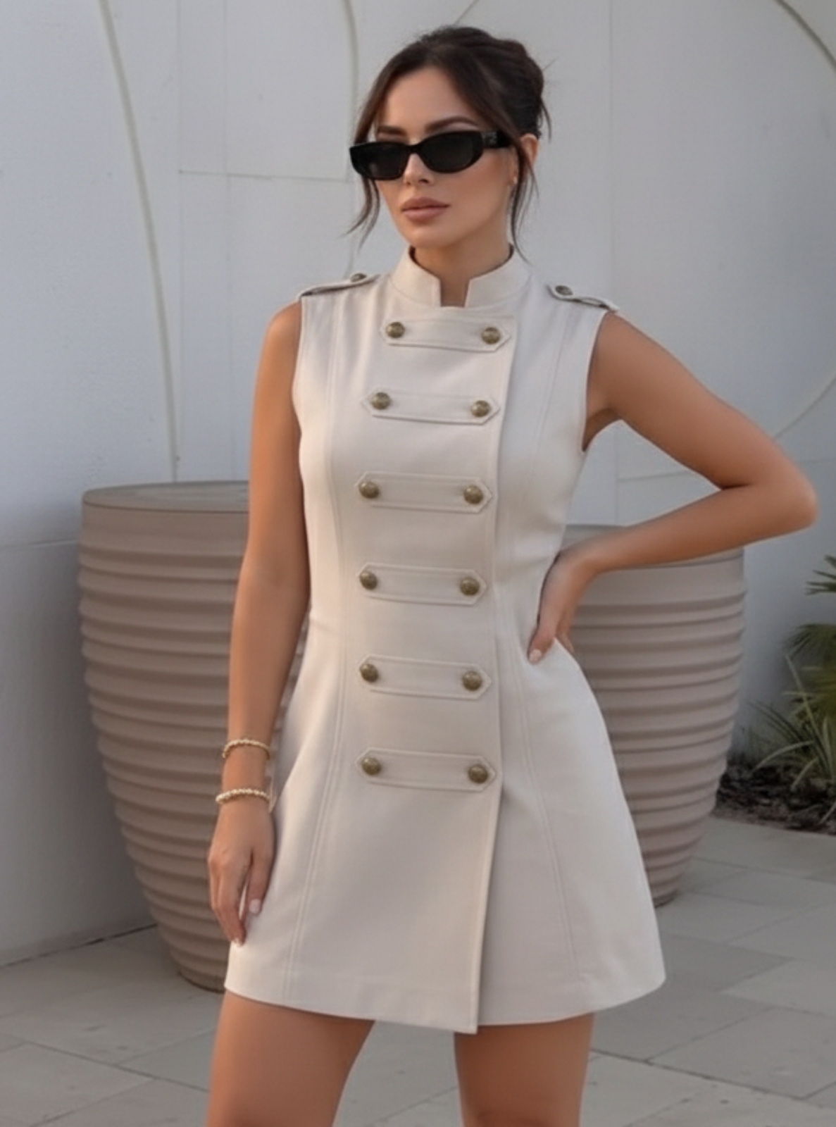 Woman wearing a beige sleeveless dress with button details against a white wall.