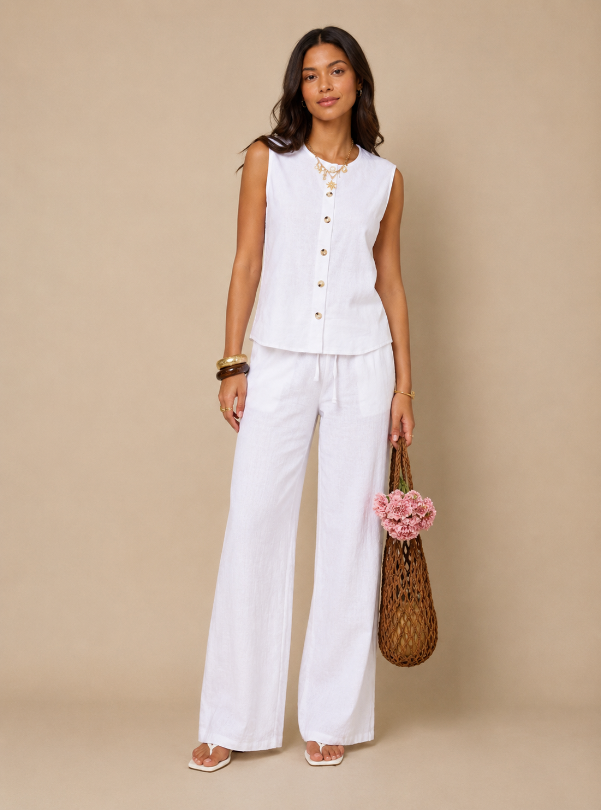A woman models a white sleeveless button-front vest and matching white wide-leg pants against a solid beige studio background. She is accessorized with gold jewelry, a woven brown bag, and a small pink flower accent.