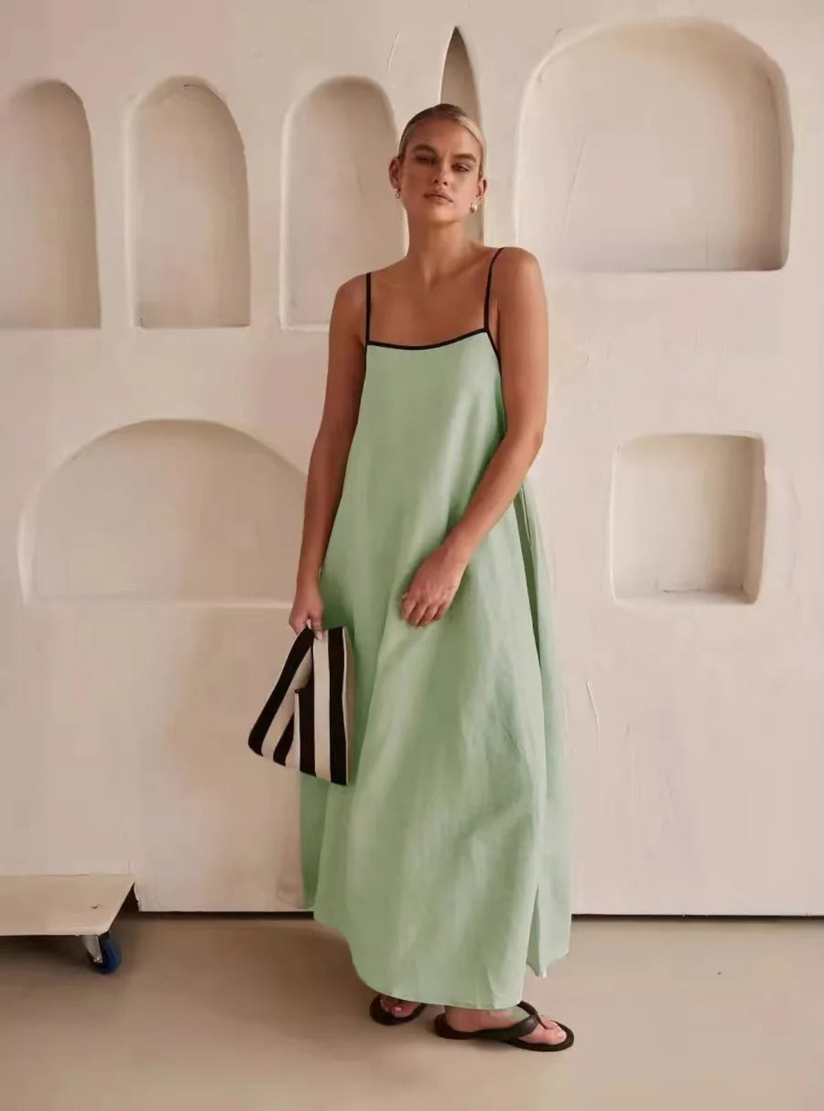 Woman in a light green slip dress holding a black and white bag against a neutral wall.