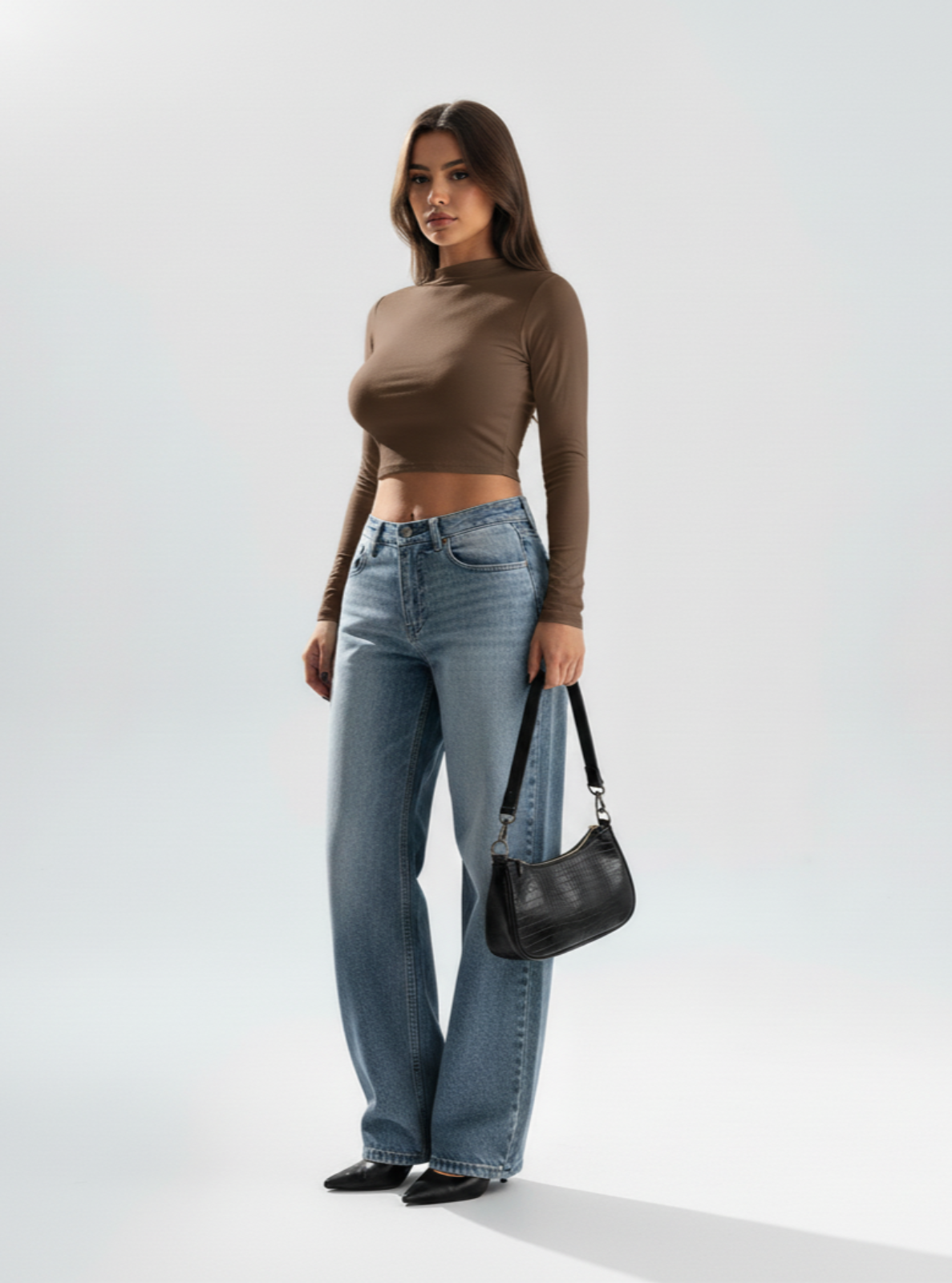 Woman wearing a brown long-sleeve top and blue jeans, holding a black handbag on a white background