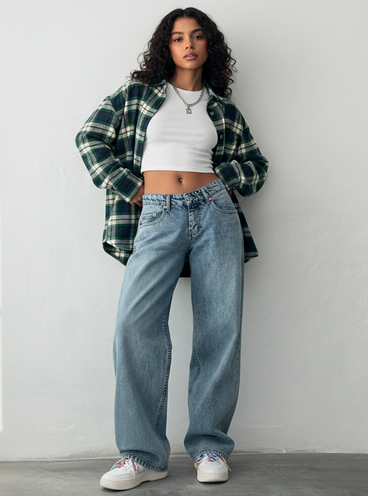 Woman wearing a plaid shirt, white crop top, and blue jeans against a white wall.