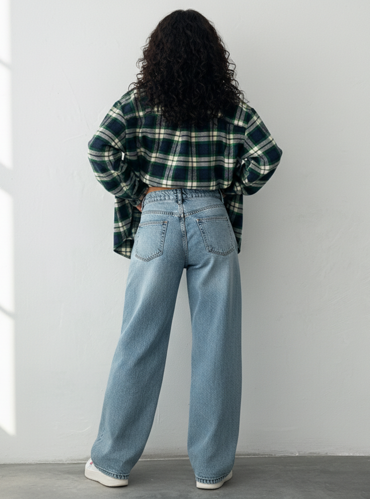 Person wearing a plaid shirt and blue jeans standing against a white wall.