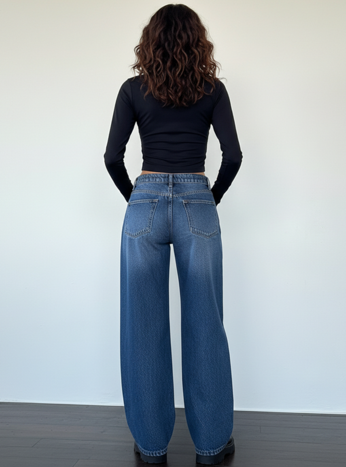 Person wearing blue jeans and a black top standing against a plain white wall.