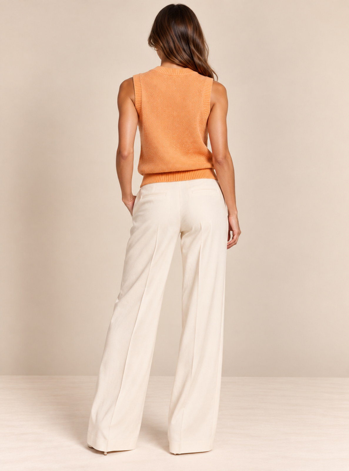 A woman shown from the back wearing a sleeveless orange knit top and off-white wide-leg trousers, standing against a plain beige studio backdrop.