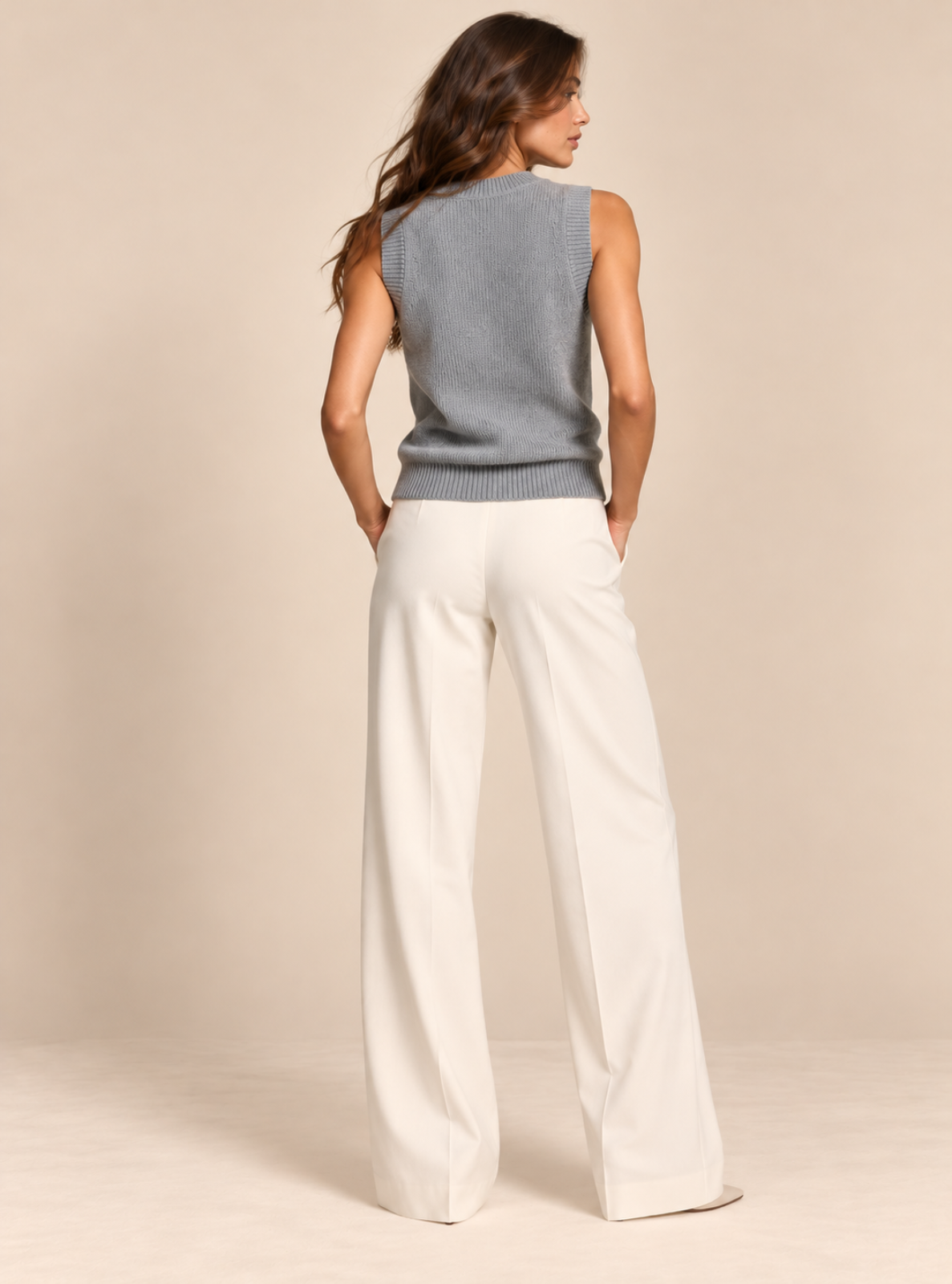 A woman shown from the back wearing a sleeveless light gray knit top and off-white wide-leg trousers, standing against a plain beige studio background.