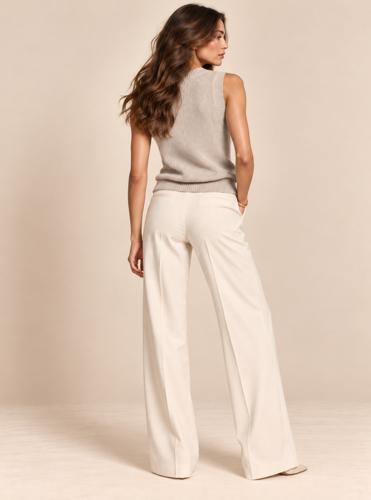 A woman shown from the back wearing a light gray sleeveless knit top and off-white wide-leg trousers, standing against a plain beige studio backdrop.