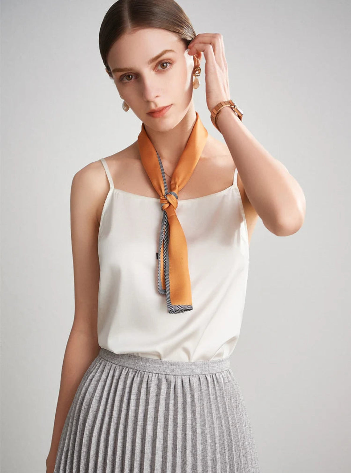 Woman wearing a white top and gray pleated skirt with an orange scarf around her neck on a light gray background