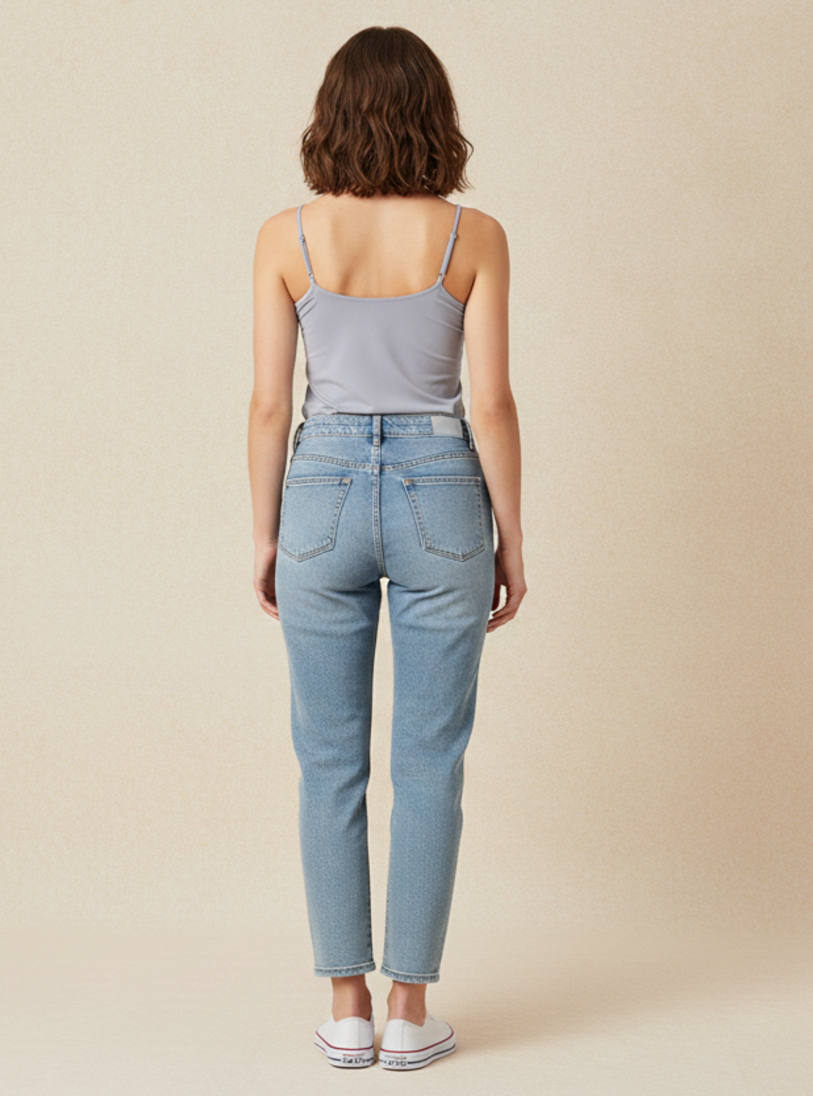 Woman wearing light blue jeans and a gray tank top on a beige background