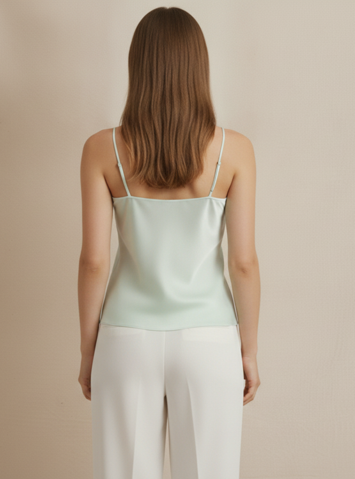 Woman wearing a light green satin camisole and white pants against a beige background