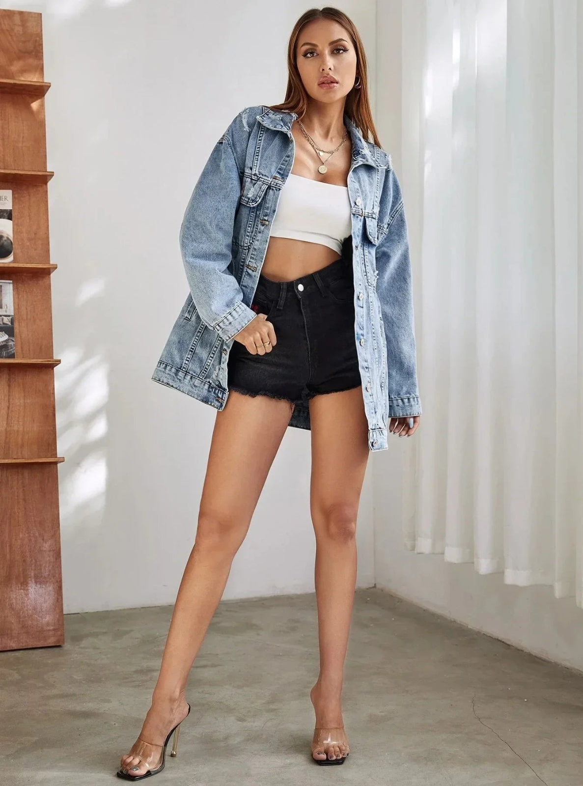 Woman wearing a denim jacket, white crop top, and black shorts in a minimal indoor setting.
