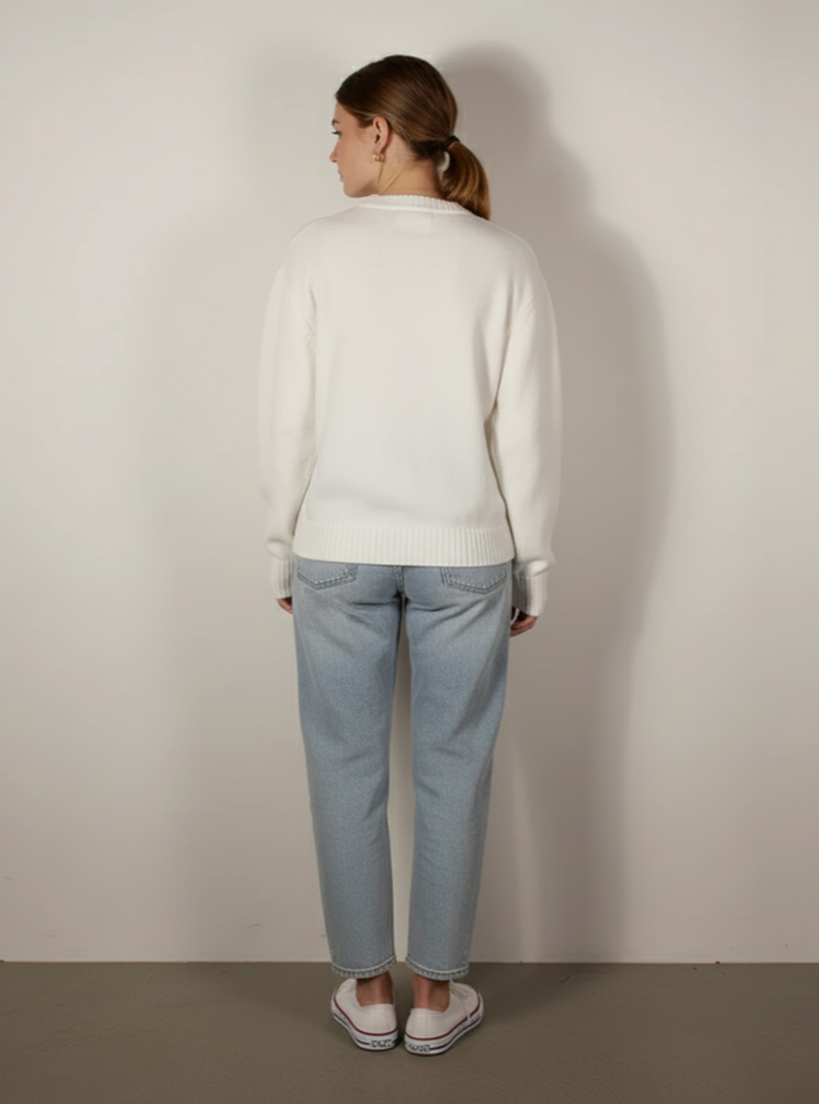 Person wearing a white sweater and light blue jeans against a plain wall.