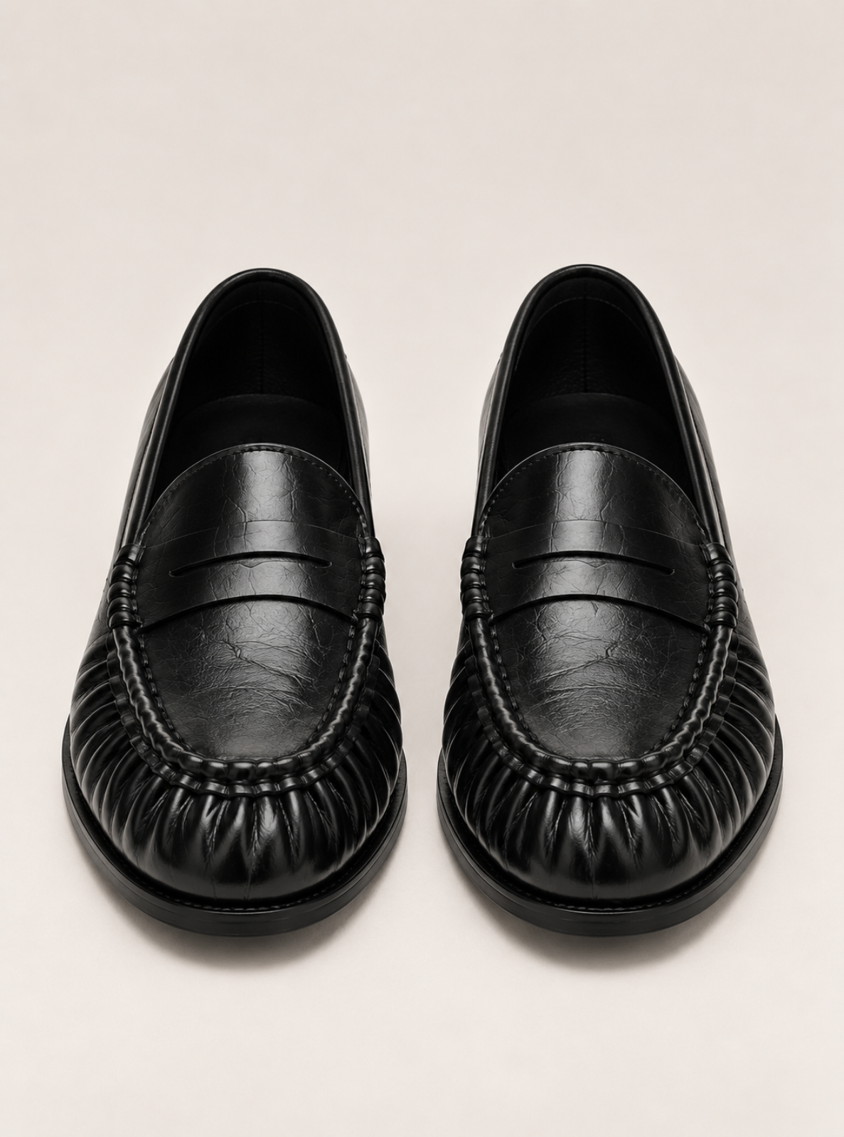 A pair of black leather penny loafers with a textured, pebbled finish and pleated detailing on the toe, shown from a top-down view on a light beige background.