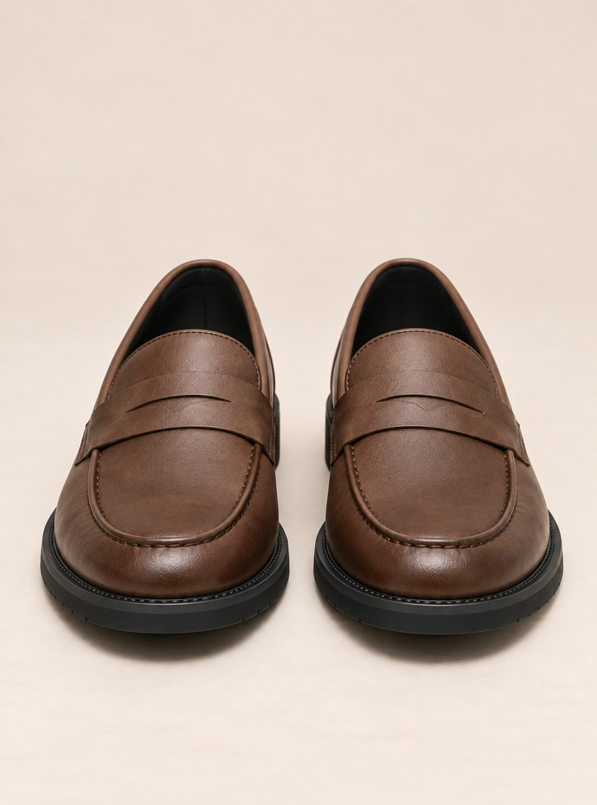 A pair of brown leather penny loafers with black soles, photographed from a front-facing perspective on a light beige background.
