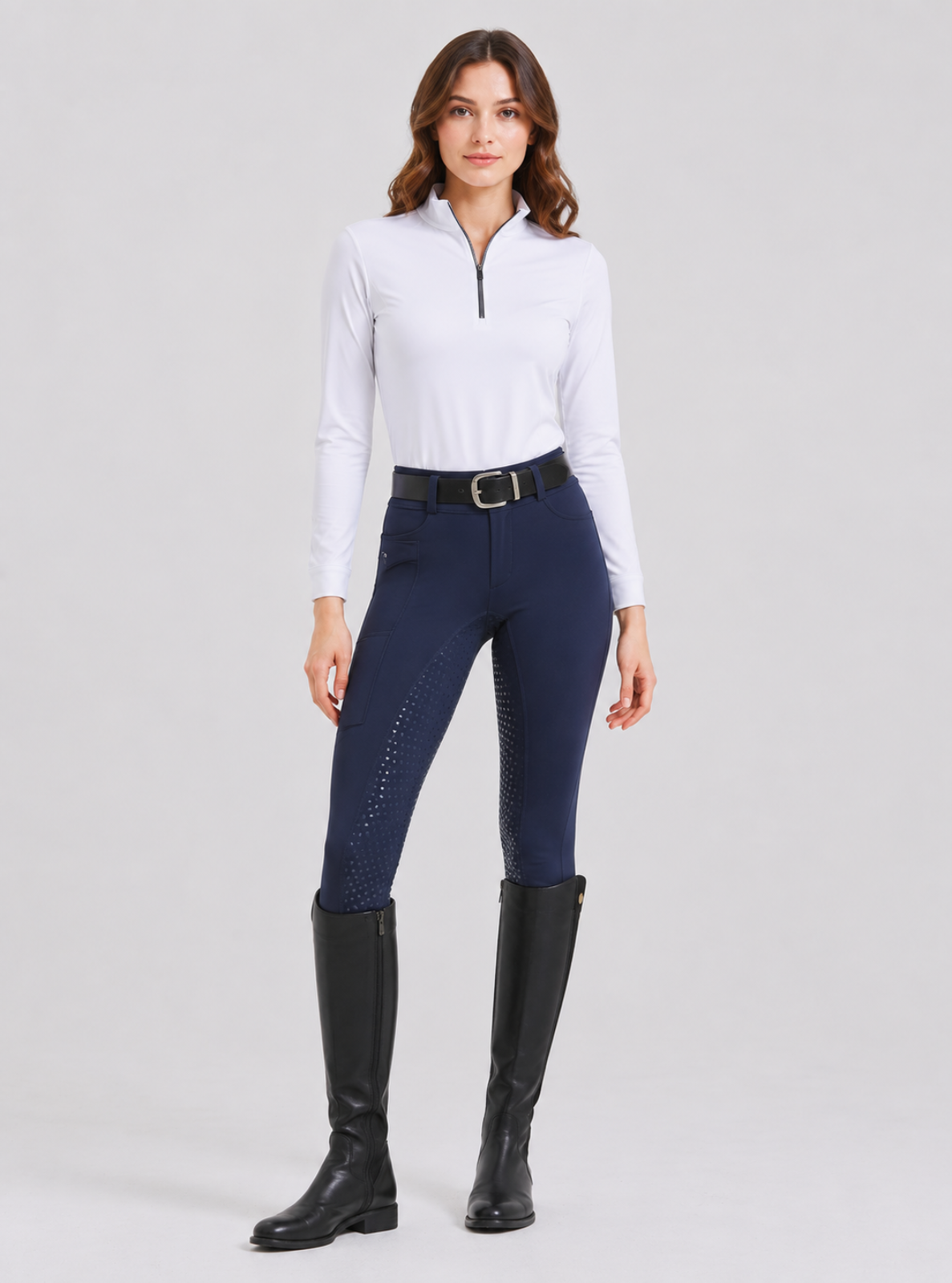 A woman modeling a white long-sleeve quarter-zip performance shirt, navy blue riding breeches with subtle side stitching, a black leather belt, and black riding boots, against a light gray studio background.