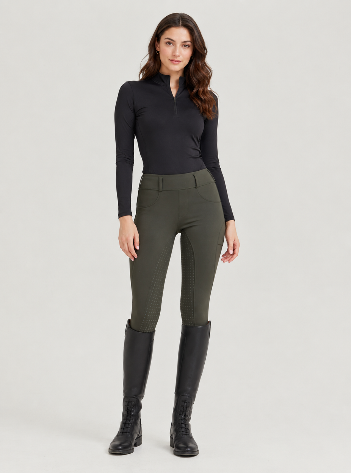A woman modeling equestrian apparel, featuring dark olive green riding breeches with textured paneling down the leg, a black long-sleeve half-zip top, and black riding boots, set against a solid light gray studio background.