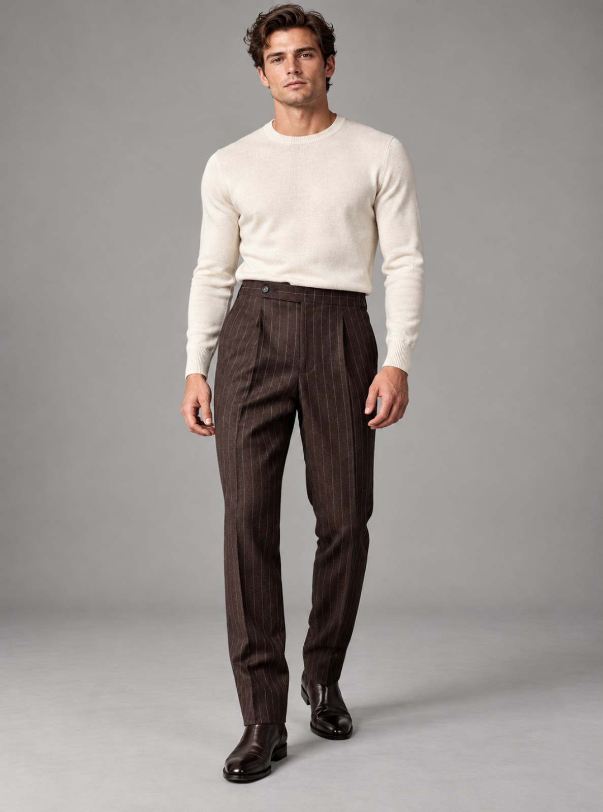 A male model wearing a cream knit sweater and dark brown pinstriped trousers, standing against a neutral gray studio background.