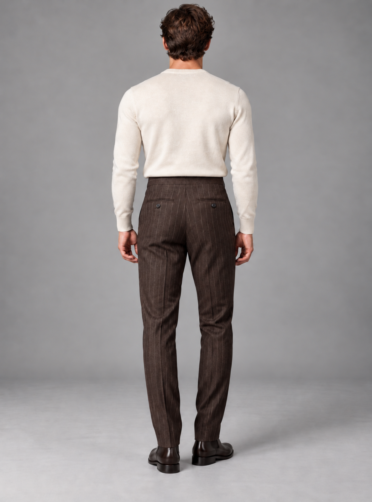 A man viewed from behind wearing a cream knit sweater and dark brown pinstriped trousers, standing against a neutral gray studio background.
