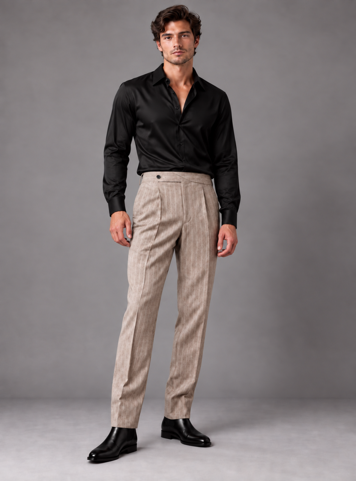 A man modeling beige pinstriped trousers with a black long-sleeve button-down shirt and black dress shoes, standing against a neutral gray studio background.