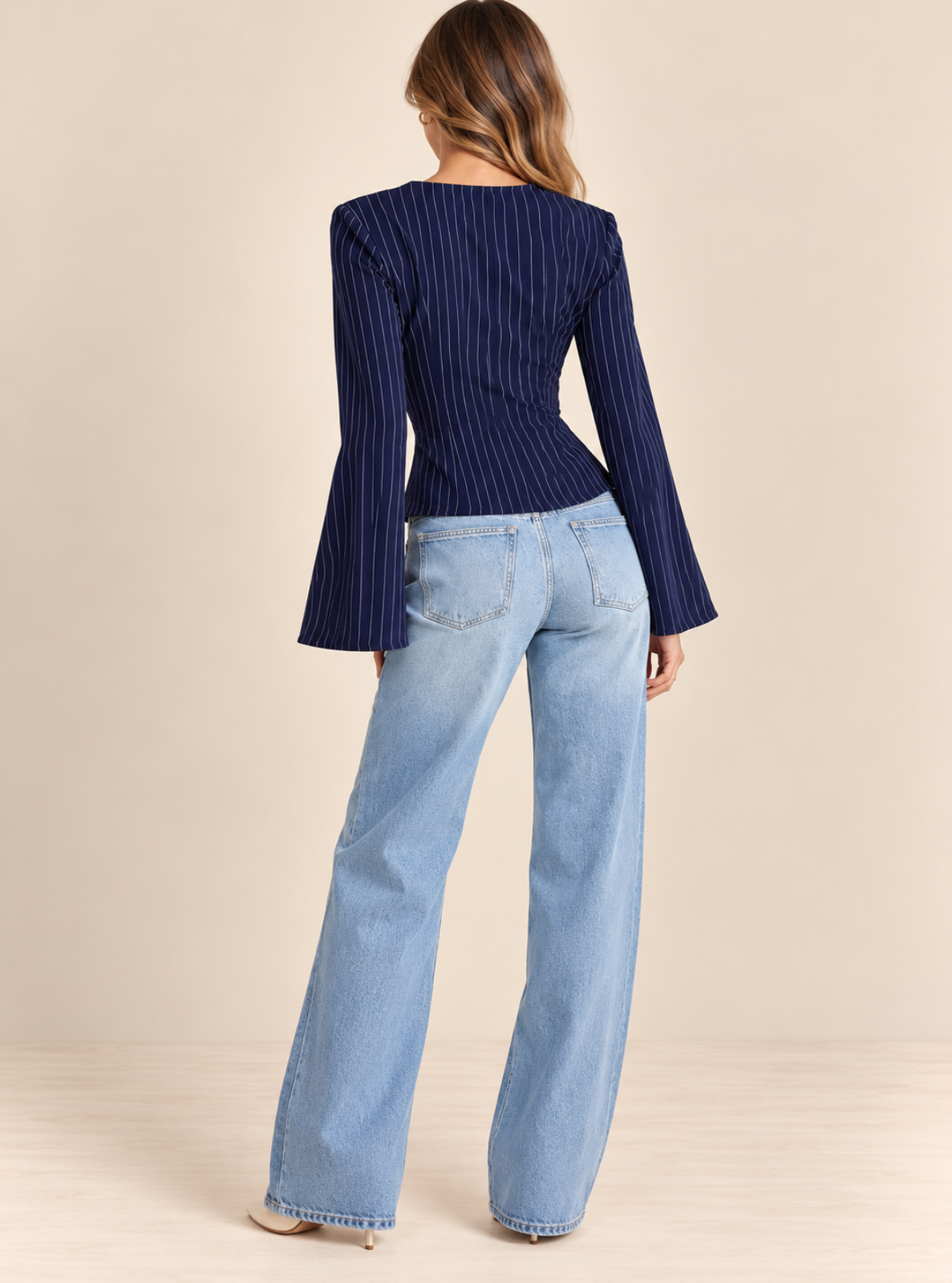 A woman shown from the back wearing a navy blue long-sleeve top with vertical pinstripes and light blue wide-leg jeans, standing against a plain beige studio background.
