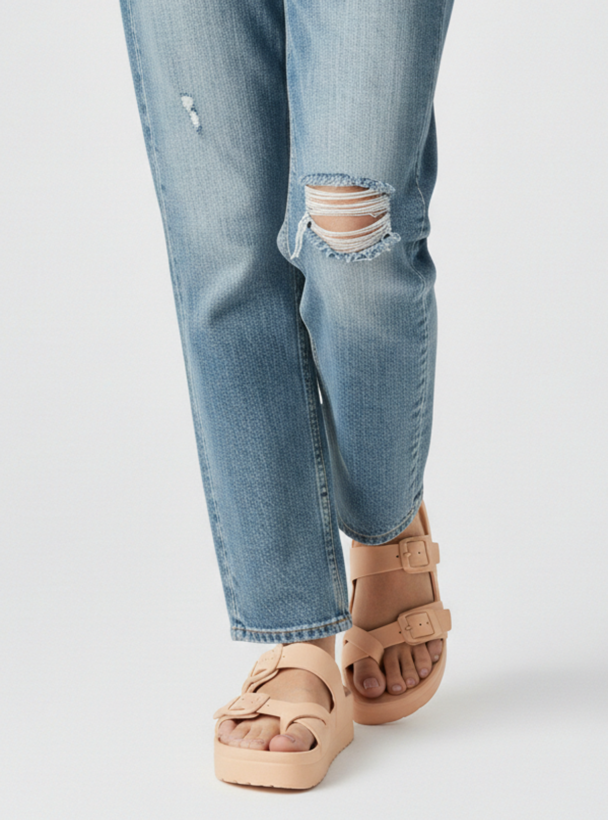 Person wearing light blue ripped jeans and beige sandals on a white background
