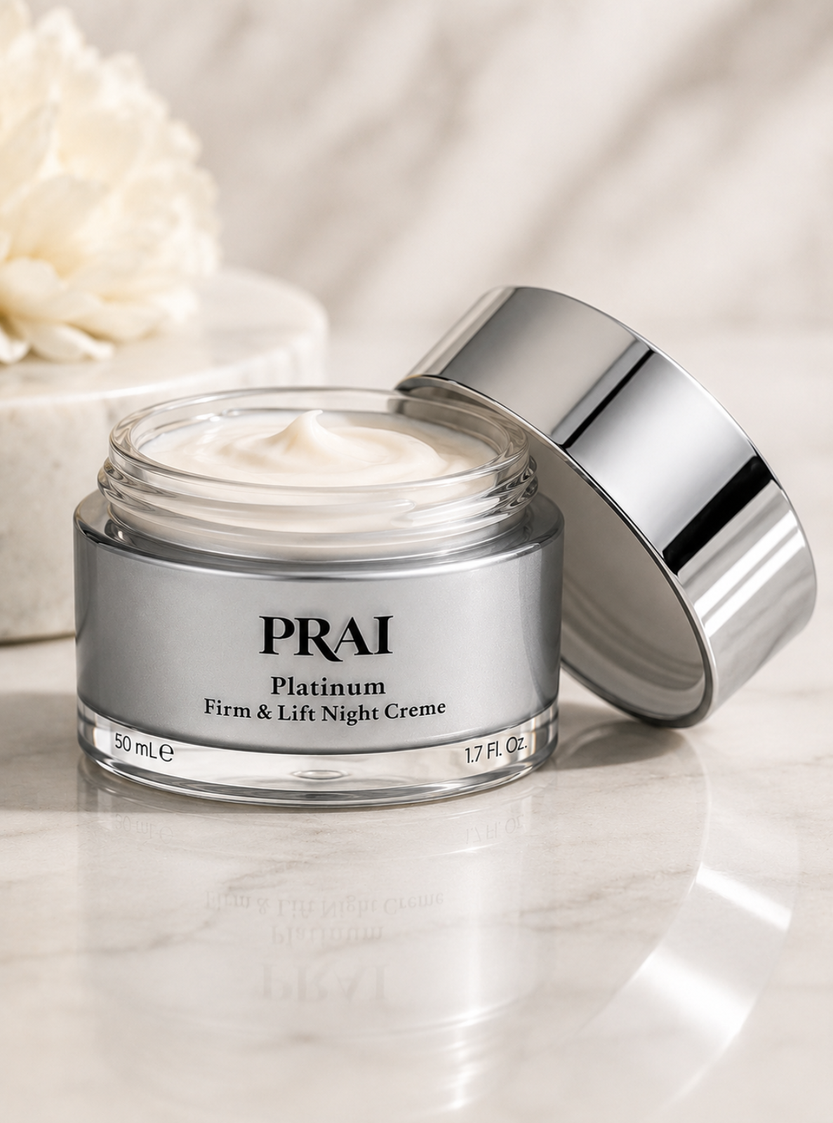PRAI Platinum Firm & Lift Night Creme in a clear glass jar with a reflective silver lid, set on a light marble surface with a soft-focus white flower in the background.